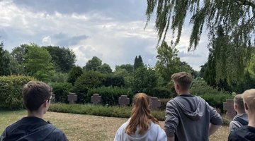 Eine Gruppe junger Menschen steht auf einer Wiese vor mehreren dunklen Steinkreuzen auf einem Friedhof. Die Szene spielt an einem bewölkten Tag, umgeben von grünen Bäumen und Büschen