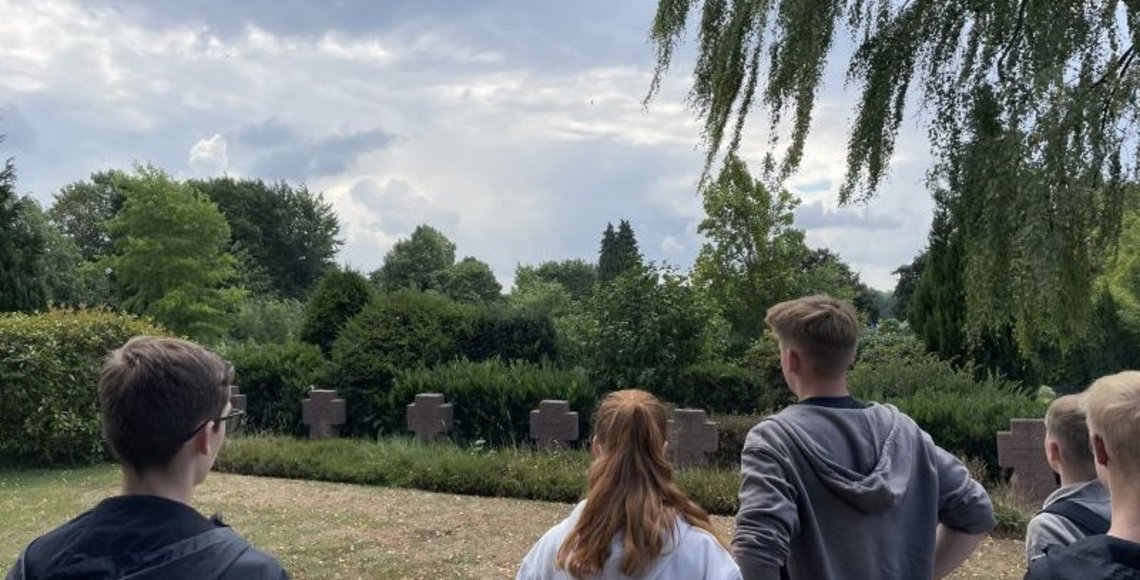 Eine Gruppe junger Menschen steht auf einer Wiese vor mehreren dunklen Steinkreuzen auf einem Friedhof. Die Szene spielt an einem bewölkten Tag, umgeben von grünen Bäumen und Büschen