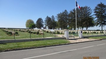 Das Foto zeigt diesmal eine andere (französische) Kriegsgräberstätte. Hinter der Straße sieht man das Eingangstor sowie die französische Flagge am Fahnenmast und dahinter viele Reihen von hellen Kreuzen auf der Anlage.