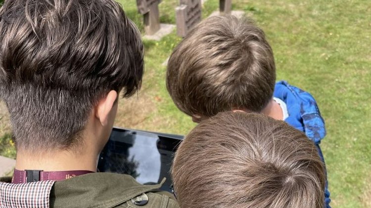 Drei Jungen stehen dicht beieinander auf einem Friedhof und blicken auf ein Tablet. Im Hintergrund sind mehrere Grabkreuze und gepflegte Gräber zu sehen. Die Sonne scheint, und die Szene spielt sich auf einer Rasenfläche ab