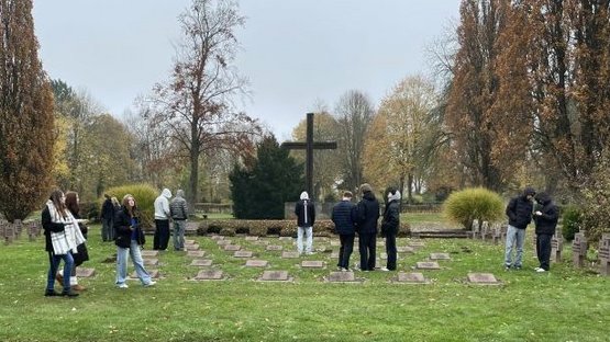 Schüler und Schülerinnen stehen in Gruppen auf dem Kriegsgräberfeld und bearbeiten Aufgaben am Tablet.