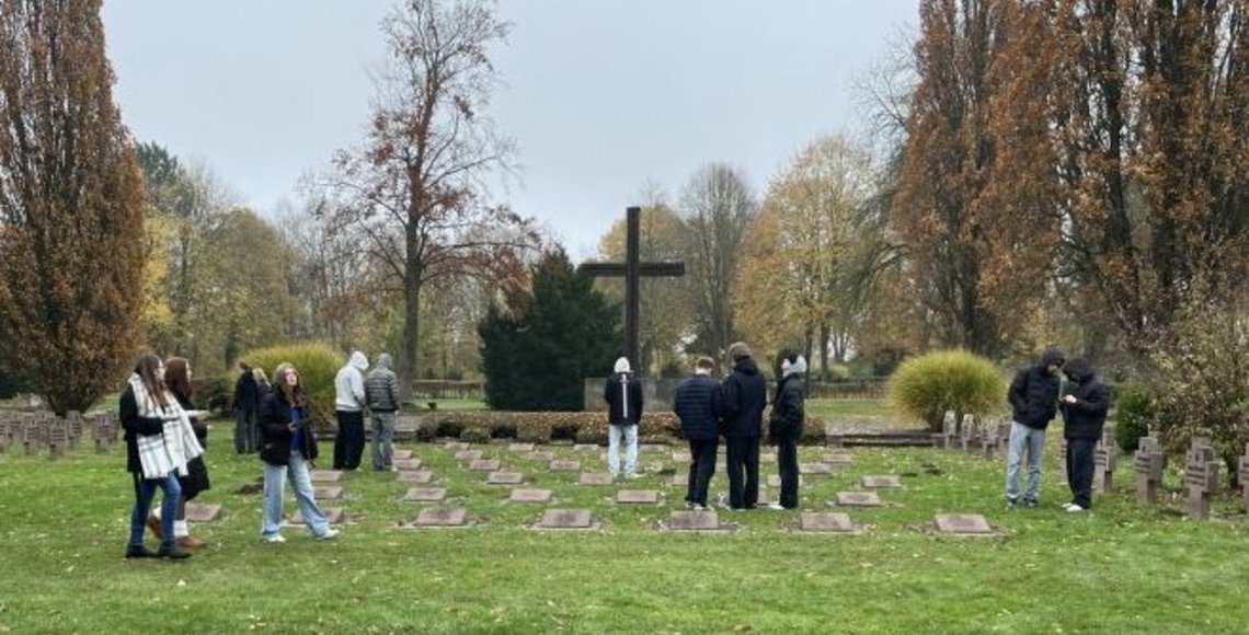 Schüler und Schülerinnen stehen in Gruppen auf dem Kriegsgräberfeld und bearbeiten Aufgaben am Tablet.